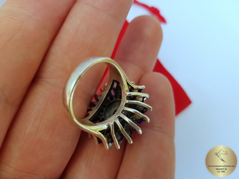 May include: A silver ring with a large purple gemstone surrounded by smaller purple gemstones. The ring is set in a silver band with a delicate design.