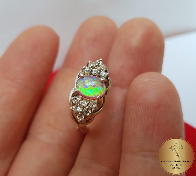 May include: A silver ring with a large oval opal gemstone surrounded by smaller clear gemstones. The opal has a rainbow of colors, including green, pink, and blue.