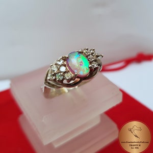 May include: A silver ring with an oval opal gemstone and smaller clear gemstones set around it. The ring is on a white display stand.