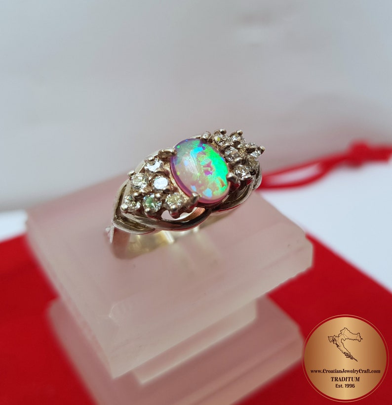 May include: A silver ring with an oval opal gemstone and smaller clear gemstones set around it. The ring is on a white display stand.