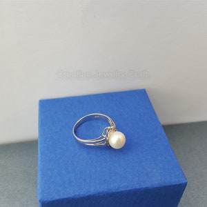 May include: A silver ring with a single white pearl set in a delicate design. The ring is displayed on a blue box.