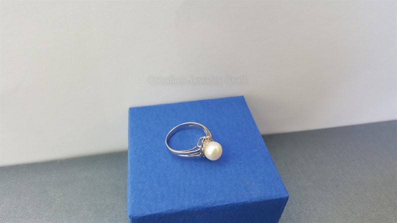 May include: A silver ring with a single white pearl set in a delicate design. The ring is displayed on a blue box.