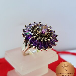 May include: A silver ring with a large purple gemstone surrounded by smaller purple gemstones. The ring is set with marcasite stones and has a floral design.