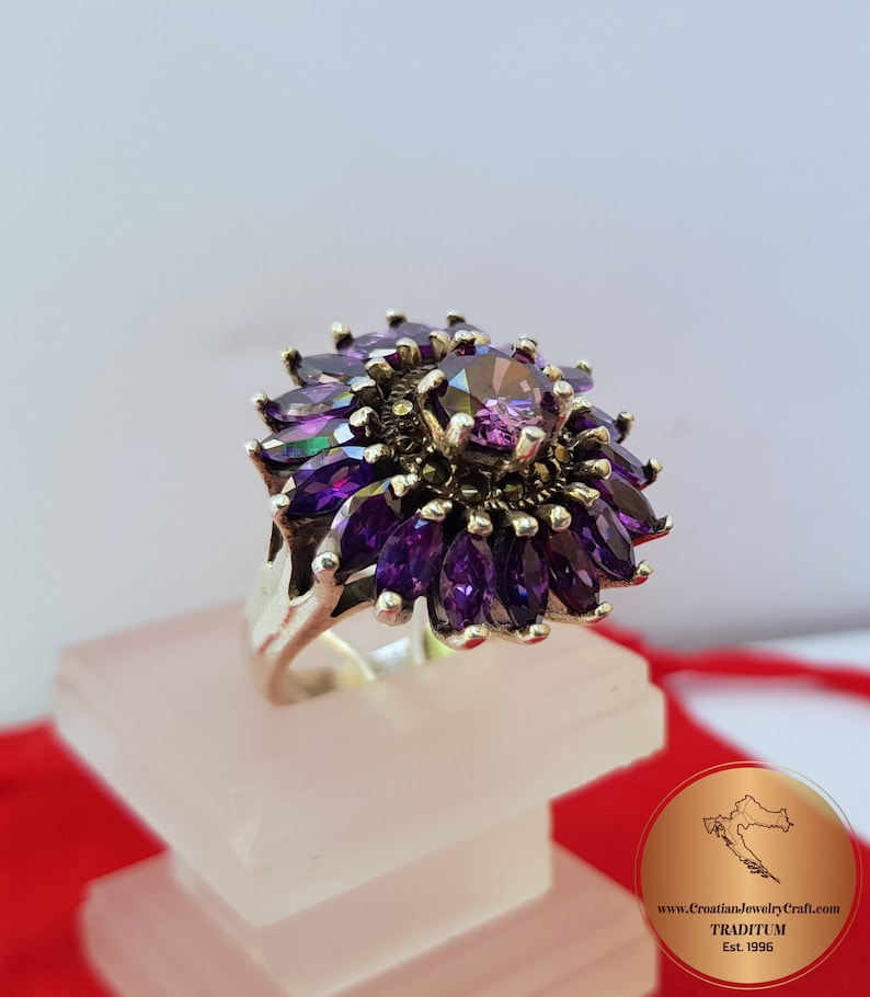 May include: A silver ring with a large purple gemstone surrounded by smaller purple gemstones. The ring is set with marcasite stones and has a floral design.