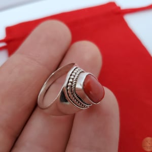 Może przedstawiać: Srebrny pierścionek z czerwonym koralem osadzonym w oprawie typu bezel. Pierścionek ma zdobienie w postaci koralik&oacute;w wok&oacute;ł kamienia.