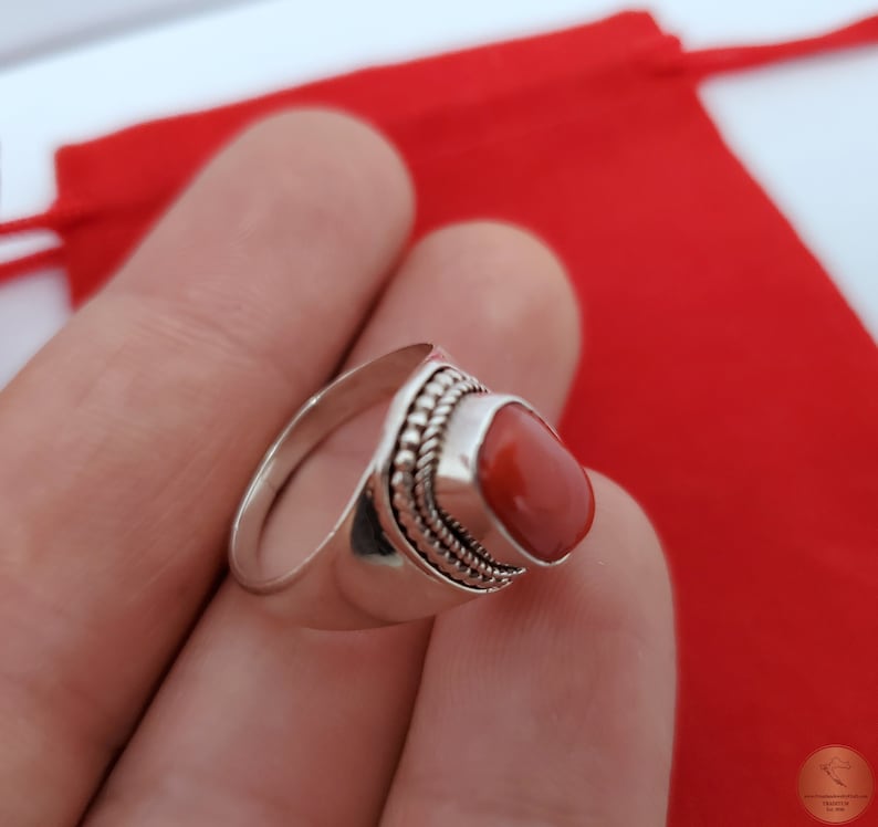 Może przedstawiać: Srebrny pierścionek z czerwonym koralem osadzonym w oprawie typu bezel. Pierścionek ma zdobienie w postaci koralik&oacute;w wok&oacute;ł kamienia.