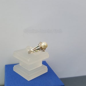 May include: A silver ring with a white pearl set in a delicate floral design. The ring is displayed on a clear acrylic stand.