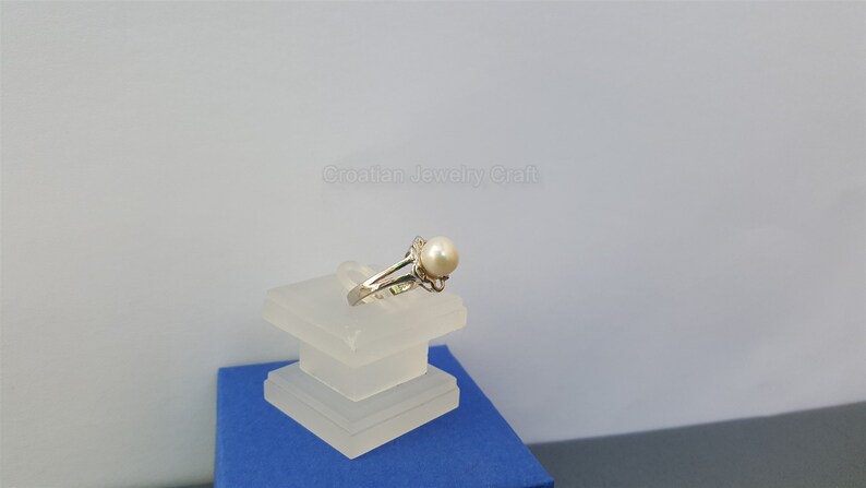 May include: A silver ring with a white pearl set in a delicate floral design. The ring is displayed on a clear acrylic stand.