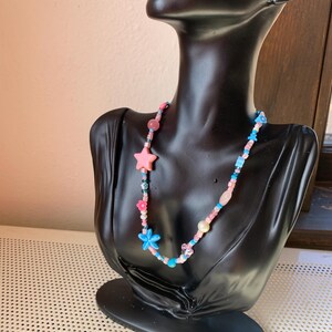 Op de afbeelding: Een kleurrijke kralenketting met roze ster- en bloembedels, blauwe en roze kralen en diverse andere decoratieve kralen. De ketting wordt getoond op een zwarte mannequin buste.