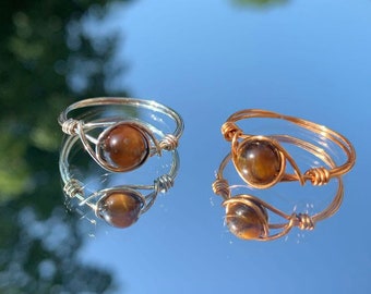 May include: Two wire-wrapped rings with a polished, round, brown gemstone. One ring is silver-toned, the other is gold-toned. The rings are displayed on a reflective surface with a blurred background of blue sky and green foliage.