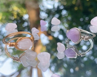May include: Two wire-wrapped rings with gemstones. One ring has a gold-colored wire and a pink gemstone. The other ring has a silver-colored wire and a purple gemstone. Small gemstones are scattered around the rings.
