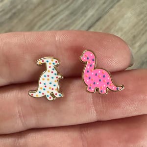 May include: Two dinosaur-shaped enamel pins. One is white with colorful polka dots, the other is pink with pink polka dots. Both pins have gold-colored outlines.