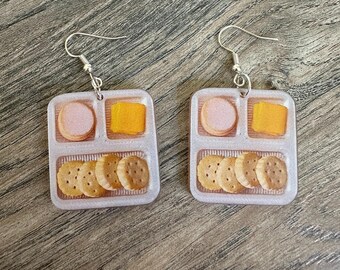 May include: A pair of novelty earrings featuring a miniature, stylized lunch tray design. Each earring showcases a pink circle, a yellow square, and several brown crackers, all set within a clear, rectangular tray. The earrings have silver-colored fish hook closures.