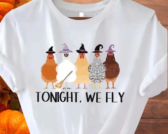 May include: White t-shirt featuring a whimsical Halloween design. Five chickens, each adorned with a witch hat, stand in a row. The text "TONIGHT, WE FLY" is printed below the chickens in black. The shirt is hanging on a wooden hanger.