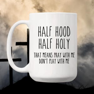 May include: White ceramic mug with the text "HALF HOOD HALF HOLY THAT MEANS PRAY WITH ME DON'T PLAY WITH ME" in black. The mug has a curved handle and is set against a cloudy sky background with silhouettes of crosses.