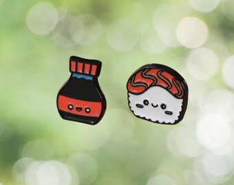 Könnte beinhalten: Ein Paar Emaille-Ohrringe zum Thema Sushi. Ein Ohrring ist eine schwarze Sojasoßenflasche mit roten und blauen Akzenten. Der andere ist eine Sushi-Rolle mit roten, weißen und schwarzen Details. Die Ohrringe haben ein niedliches, cartoonhaftes Design.