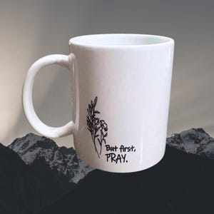 May include: White ceramic mug with a curved handle. The mug features a black floral design and the text "But first, PRAY." The background shows a mountain range.