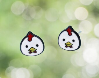 May include: A pair of chicken head stud earrings. Each earring features a white head with a black outline, a red comb, and a yellow beak. The earrings are set against a blurred green background, creating a playful and quirky accessory.