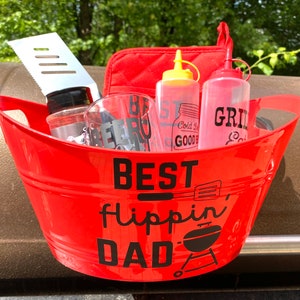 Pode incluir: Balde de metal vermelho com alças pretas cheio de acessórios para grelhar. O balde tem o texto "BEST Flippin' DAD" impresso no lado. Os acessórios incluem uma espátula, duas garrafas de vidro, duas garrafas de pressão e um pegador de panela vermelho e preto.