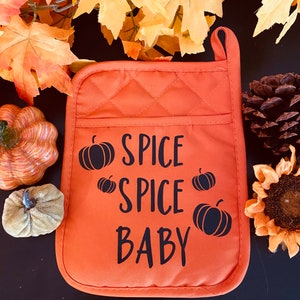 May include: An orange quilted pot holder with the text "SPICE SPICE BABY" and black pumpkin graphics. The pot holder has a loop for hanging and is surrounded by autumn-themed decorations, including leaves and pumpkins.