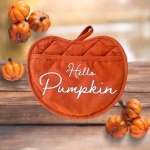 Può includere: Presa pentola imbottita di colore arancione a forma di mela con il testo "Hello Pumpkin" in bianco. La presa pentola è circondata da piccole zucche arancioni.