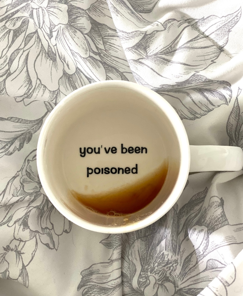 Peut inclure: Une tasse en c&eacute;ramique blanche avec le texte "you've been poisoned" imprim&eacute; &agrave; l'int&eacute;rieur. La tasse est remplie d'un liquide brun.