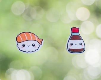 Könnte beinhalten: Zwei Aufkleber im Cartoon-Stil: einer mit Sushi mit orangefarbenem Lachs und einer weißen Reisbasis und ein weiterer mit einer Sojasoßenflasche mit rotem Deckel. Beide haben lächelnde Gesichter und schwarze Umrisse.