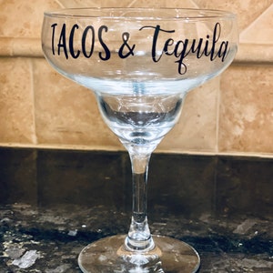 May include: Clear margarita glass with the words "TACOS & Tequila" in black script. The glass has a wide bowl, a stepped base, and a long stem. The glass is on a dark countertop.