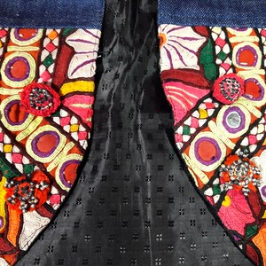 May include: A close-up of a denim jacket with colorful embroidered patches and beaded embellishments. The patches feature geometric patterns, flowers, and other designs. The jacket is lined with black fabric.