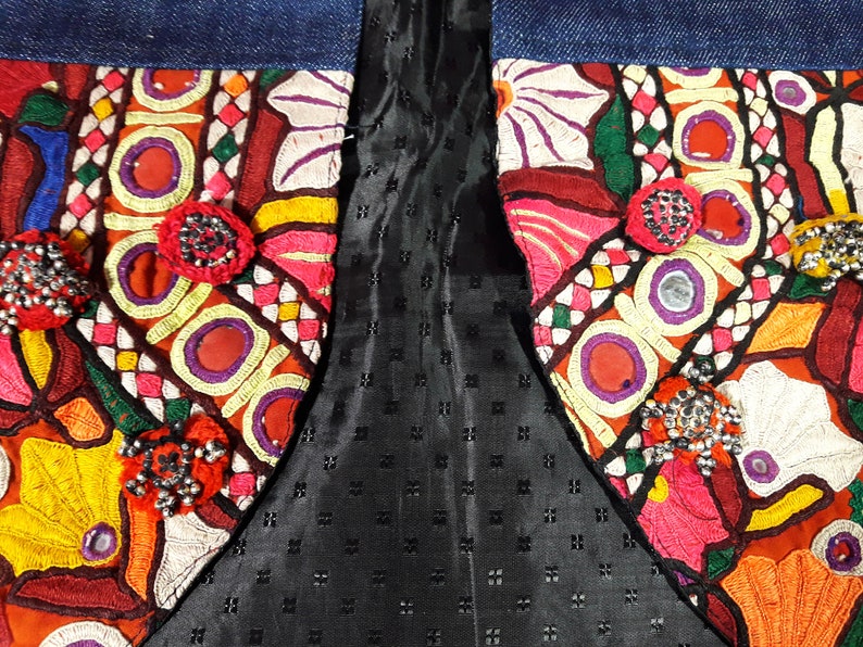 May include: A close-up of a denim jacket with colorful embroidered patches and beaded embellishments. The patches feature geometric patterns, flowers, and other designs. The jacket is lined with black fabric.