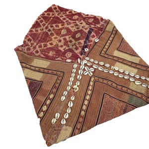 May include: Textile with geometric patterns in shades of red, brown, and beige. The fabric is adorned with white seashells, arranged in a decorative pattern. The item appears to be a shawl or wrap.