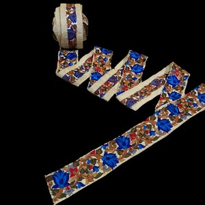 May include: A cream-colored fabric trim with blue, brown, and red floral embroidery. The trim is folded and arranged in a decorative pattern.