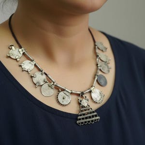 Puede incluir: Collar plateado con cordón negro. Presenta varios colgantes, incluyendo monedas redondas y formas geométricas. La joya se luce sobre una blusa azul oscuro.