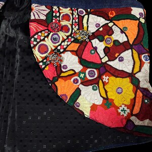 May include: A close-up of a colorful embroidered patch with a floral design. The patch is made of various fabrics and features beads and mirrors. The patch is sewn onto a denim fabric.