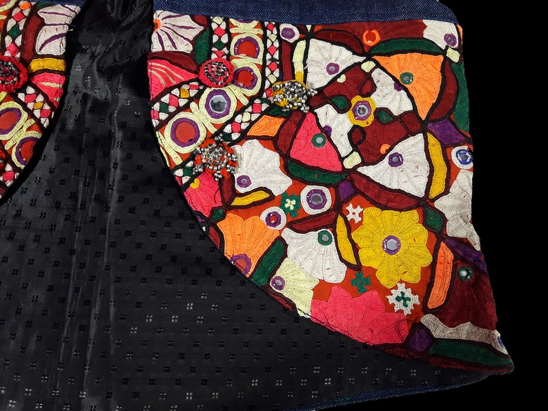 May include: A close-up of a colorful embroidered patch with a floral design. The patch is made of various fabrics and features beads and mirrors. The patch is sewn onto a denim fabric.