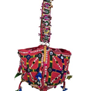 Può includere: Una borsa in tessuto rosso vivace con un motivo geometrico di linee cucite verdi e blu, specchi circolari e dettagli in conchiglia bianca. La borsa include una lunga tracolla abbinata decorata con specchi e piccoli pon-pon colorati.