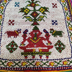 Puede incluir: Un cuadrado de tela blanca con un diseño de cuentas de colores. El diseño presenta una figura central con dos animales a cada lado, todo rodeado por un borde floral.