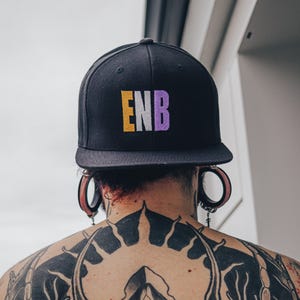 Könnte beinhalten: Eine schwarze Baseballkappe mit den Buchstaben "ENB" in Gelb, Weiß und Lila gestickt.