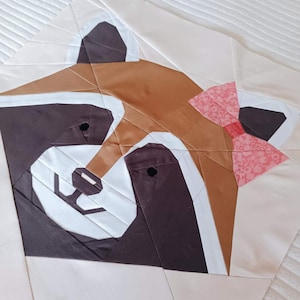 May include: A fabric quilt block featuring a stylized red panda face constructed from geometric pieces. The design uses shades of brown, dark gray, and white fabric, with a small pink floral patterned bow attached to the side of the head.