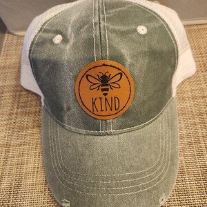 May include: A distressed green trucker hat with a white mesh back. A circular brown patch features a bee illustration and the word "KIND". The hat has a curved brim and a distressed appearance.