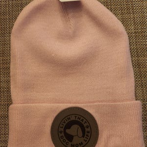 May include: A light pink beanie with a folded brim. A circular patch on the brim reads "Livin' That Dog Mom Life" with a dog silhouette. A price tag is attached.