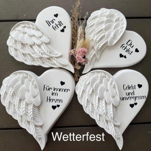 Könnte beinhalten: Vier weiße, herzförmige Keramikanhänger mit Engelsflügeln. Jedes Herz trägt eine andere Botschaft in schwarzer Schrift: "Ihr fehlt", "Du fehlst", "Für immer im Herzen" und "Geliebt und unvergessen". Die Anhänger sind wetterfest.