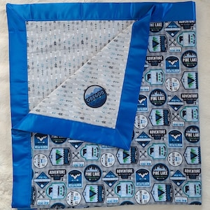 May include: A gray and blue baby blanket with a blue satin border. One side features a pattern of arrows and the other side features a pattern of camping-themed patches with the text "Pine Lake Basecamp" and "Adventure".