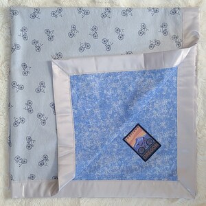 May include: A light blue baby blanket with a white satin border. The blanket has a white background with a repeating pattern of black bicycles. The blanket has a blue patch with a white mountain scene and the text "Portland Oregon".