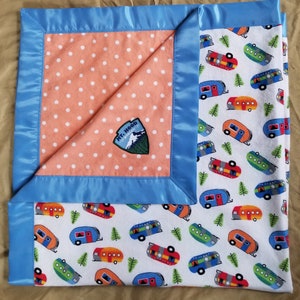 May include: A blue and white baby blanket with a peach polka dot panel and a patch that says "Mt. Hood Oregon". The other side of the blanket features a white background with a repeating pattern of colorful camper vans and green trees.