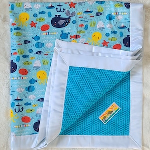 May include: A blue and white baby blanket with a sea life pattern. The blanket has a white satin border and a blue satin backing with a polka dot pattern. The blanket has a small tag that says "The Sleepy Owl".