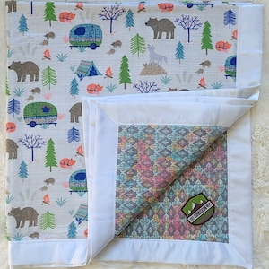 May include: A white muslin baby blanket with a woodland camping print featuring bears, foxes, and a camper van. The blanket has a white satin border and a gray and teal patterned backing with a "Lost Oregon" patch.