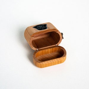 May include: A wooden box with a hinged lid, open to reveal the interior. The box is decorated with a black and white graphic of a character's face.