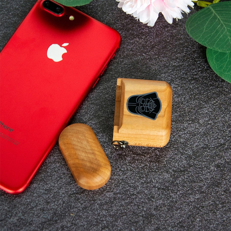 May include: A wooden case with a black Darth Vader silhouette design. The case is closed and has a small metal loop on the side. The case is sitting on a gray surface next to a red iPhone.