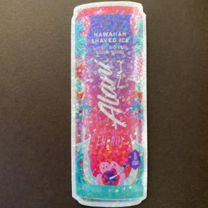 May include: A holographic sticker of a can of Aloha energy drink. The can is pink and blue with a design of palm trees and flowers. The text on the can reads "Hawaiian Shaved Ice" and "Aloha Energy".
